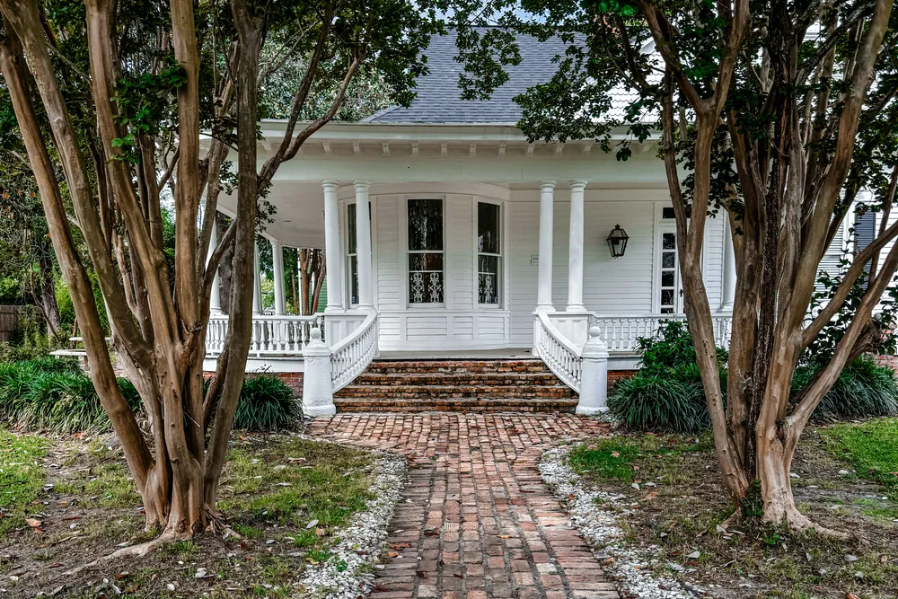 A white Louisiana cottage with a columned porch, brick path and front steps, framed by crape myrtle trunks